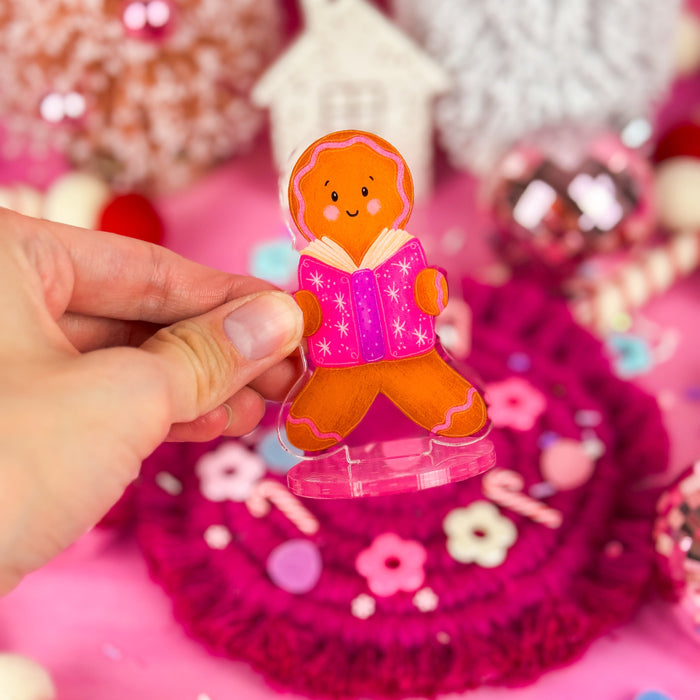 A hand holding a clear acrylic stand featuring an illustration of a gingerbread holding an open book.