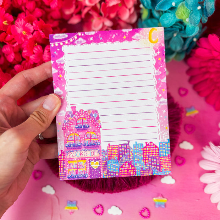 A hand holds a pink lined notepad with a colorful bookshop illustration at the bottom, surrounded by bright pink and blue flowers on a pink background.