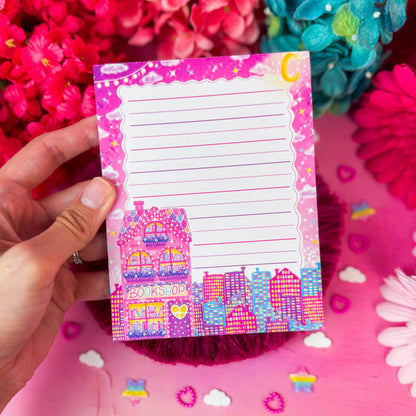 A hand holds a pink lined notepad with a colorful bookshop illustration at the bottom, surrounded by bright pink and blue flowers on a pink background.