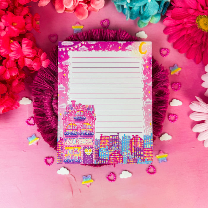 A pink lined notepad with a colorful bookshop illustration and decorative border, placed on a pink textured surface surrounded by flowers and small pastel accents.