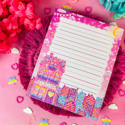 A pink lined notepad with a colorful bookshop illustration and decorative border, placed on a pink textured surface surrounded by flowers and small pastel accents.