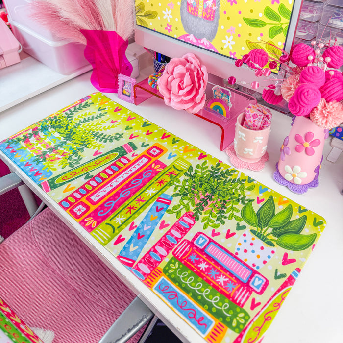 A colorful desk mat with a pattern of books, leaves, and hearts in bright green, pink, orange, and blue, displayed on a white desk next to a computer monitor and pink accessories.