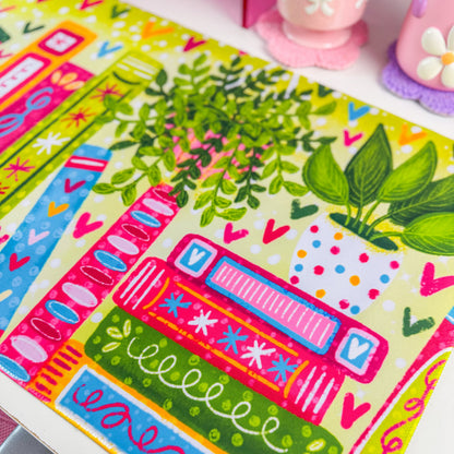 A colorful desk mat with a pattern of books, leaves, and hearts in bright green, pink, orange, and blue, displayed on a white desk next to a computer monitor and pink accessories.