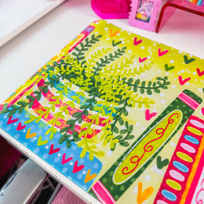A colorful desk mat with a pattern of books, leaves, and hearts in bright green, pink, orange, and blue, displayed on a white desk next to a computer monitor and pink accessories.