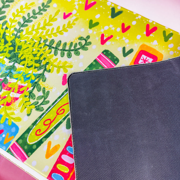 A colorful desk mat with a pattern of books, leaves, and hearts in bright green, pink, orange, and blue, displayed on a white desk next to a computer monitor and pink accessories.