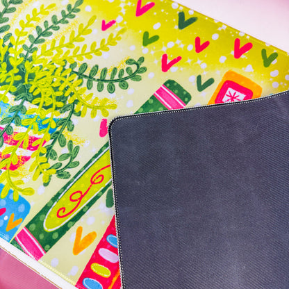 A colorful desk mat with a pattern of books, leaves, and hearts in bright green, pink, orange, and blue, displayed on a white desk next to a computer monitor and pink accessories.