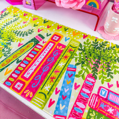 A colorful desk mat with a pattern of books, leaves, and hearts in bright green, pink, orange, and blue, displayed on a white desk next to a computer monitor and pink accessories.