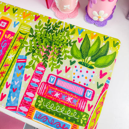 A colorful desk mat with a pattern of books, leaves, and hearts in bright green, pink, orange, and blue, displayed on a white desk next to a computer monitor and pink accessories.