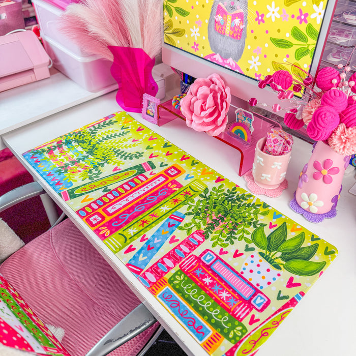 A colorful desk mat with a pattern of books, leaves, and hearts in bright green, pink, orange, and blue, displayed on a white desk next to a computer monitor and pink accessories.