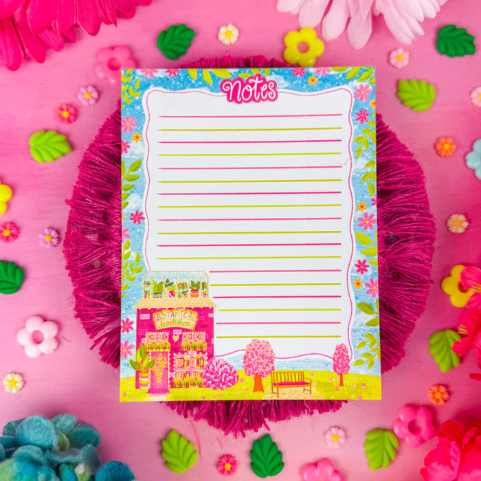 A lined notepad with the word "notes" written at the top, with a pink and orange flower border, and a botanical bookshop in the corner. Sitting on top of a bright pink background.