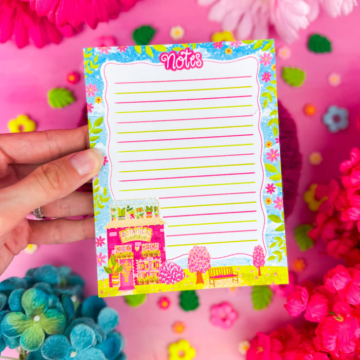 A hand holding a lined notepad with the word "notes" written at the top, with a pink and orange flower border, and a botanical bookshop in the corner. Sitting on top of a bright pink background.