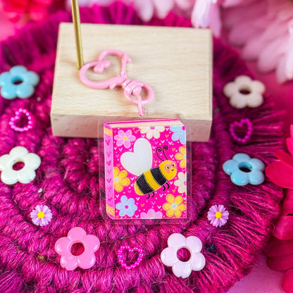 Decorative pink keychain with floral design and bee illustration on a pink background