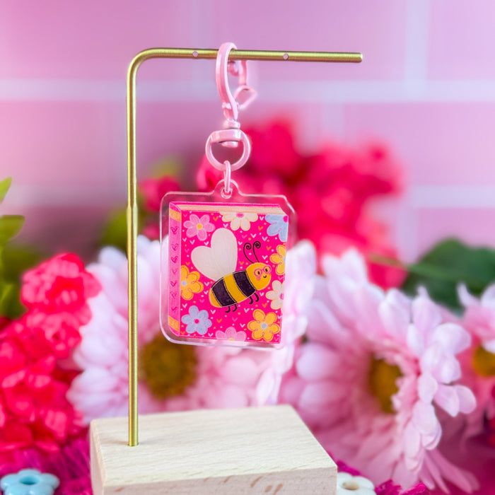Colorful bee-themed keychain on a stand with pink flowers in the background