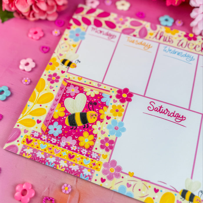 Colorful weekly planner with floral and bee design on a pink surface with flowers and decorative items.