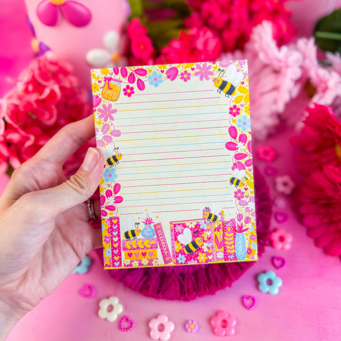 Colorful notepad with floral and bee design held by a hand against a pink floral background