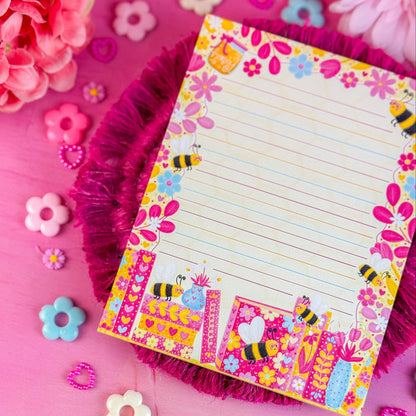 Colorful notepad with floral and bee design on a pink background with flowers and decorative items.