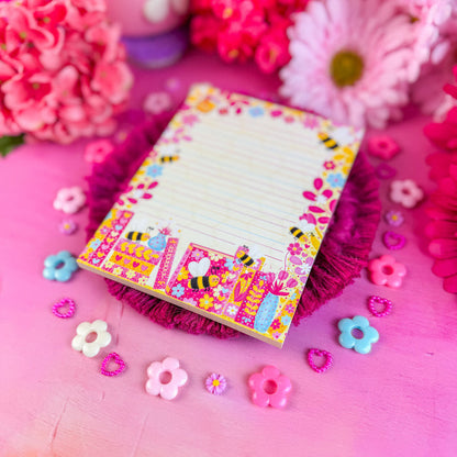 Colorful notepad with floral and bee design on a pink surface with flowers and decorative items.