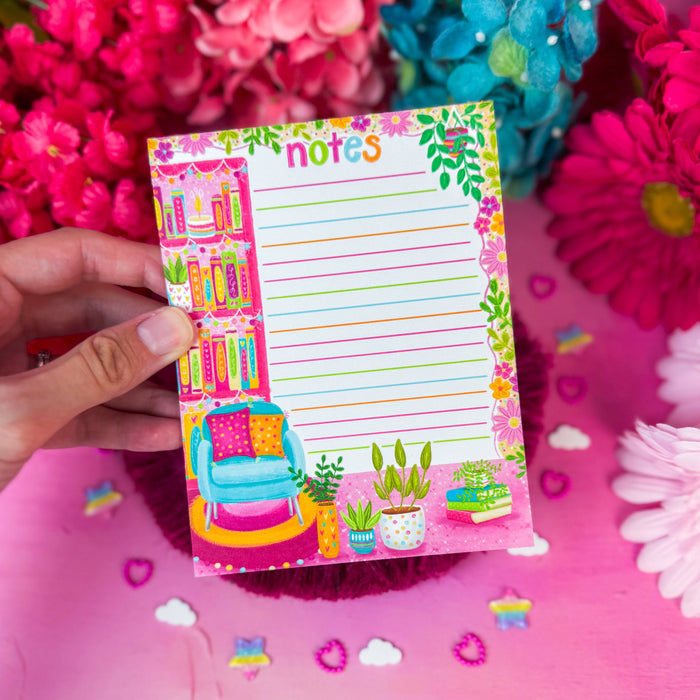 A hand holds a small colorful 'notes' notepad with a cozy library-themed illustration, surrounded by bright pink and blue flowers on a pink surface.