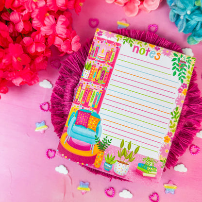 A colorful decorative notepad with lined writing space and illustrated bookshelves, flowers, and a chair, placed on a pink surface with bright pink and blue flowers around it.