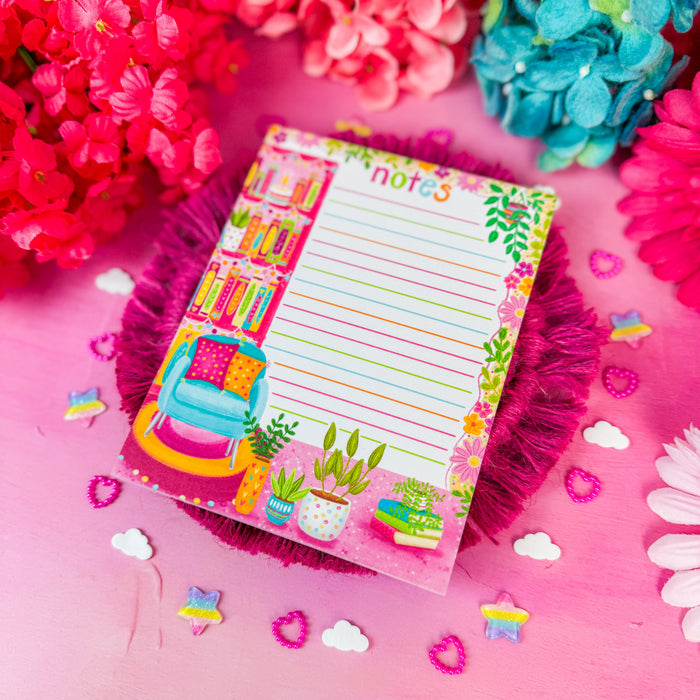 A colorful decorative notepad with lined writing space and illustrated bookshelves, flowers, and a chair, placed on a pink surface with bright pink and blue flowers around it.