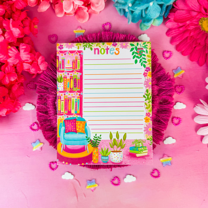 A colorful decorative notepad with lined writing space and illustrated bookshelves, flowers, and a chair, placed on a pink surface with bright pink and blue flowers around it.