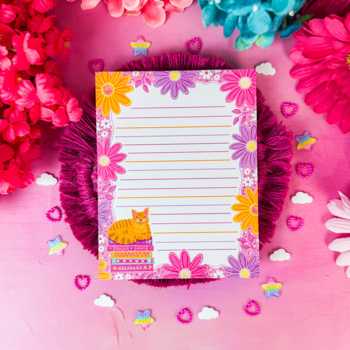 A lined notepad with a colorful floral border and a small orange cat illustration, styled on a pink background with decorative flowers and small star and cloud cutouts.