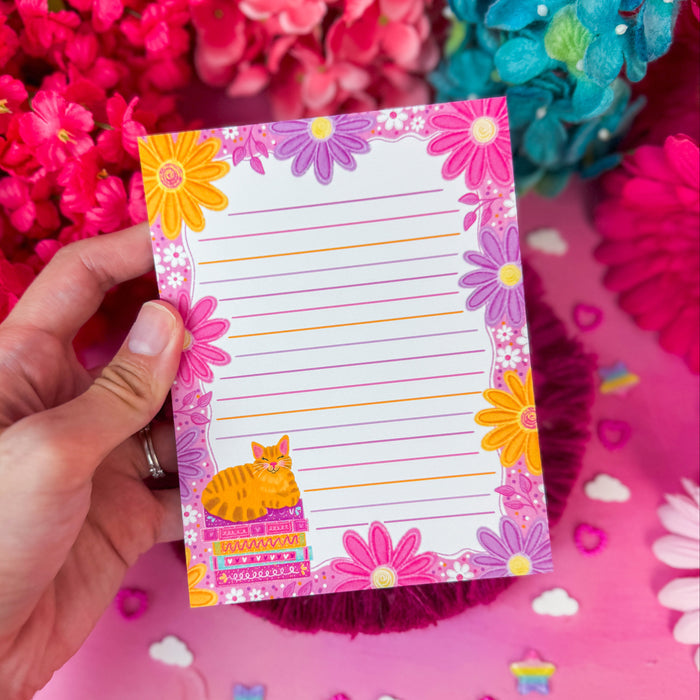 A hand holds a lined notepad with a colorful floral border and a small orange cat illustration. The background is bright pink with large flowers and small decorative items scattered around.