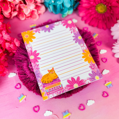A lined notepad with a colorful floral border and a small orange cat illustration, styled on a pink background with decorative flowers and small star and cloud cutouts.