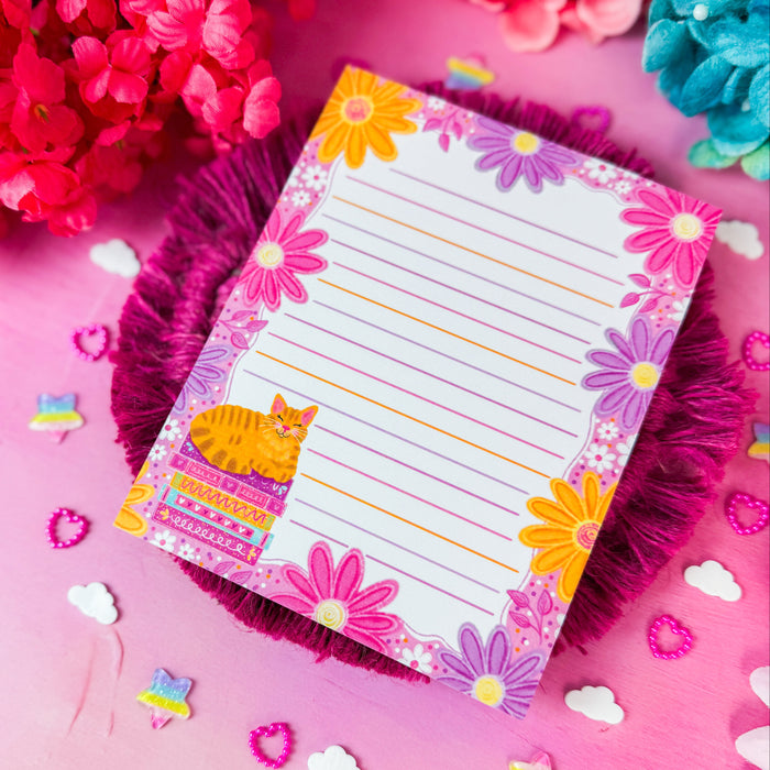 A lined notepad with a colorful floral border and a small orange cat illustration, styled on a pink background with decorative flowers and small star and cloud cutouts.