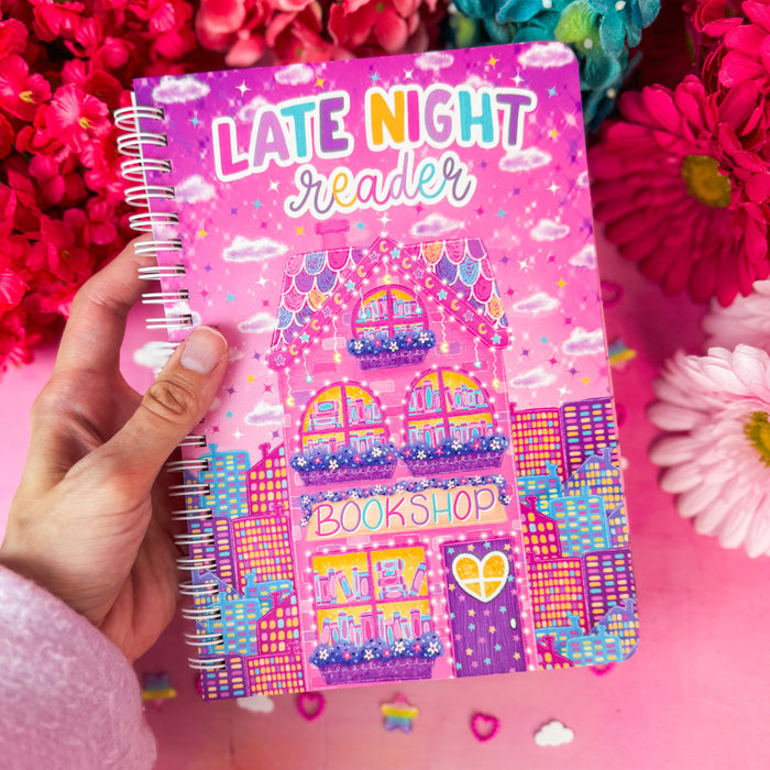 A hand holds a spiral-bound notebook titled 'LATE NIGHT reader' with a colorful bookshop illustration on the cover, set against a pink background with bright pink and blue flowers.