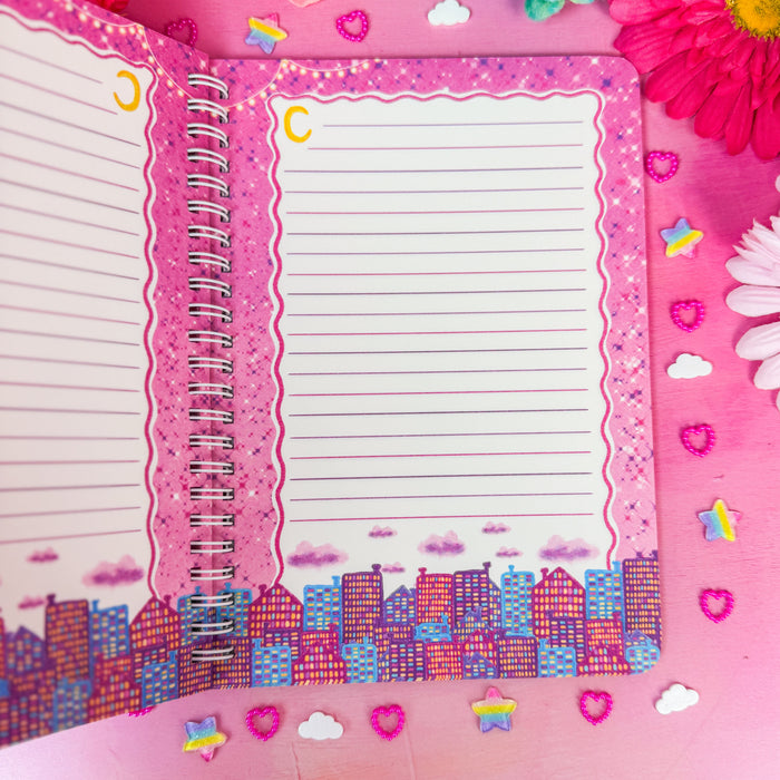 An open spiral-bound lined notebook with pink decorative borders and a colorful cityscape illustration along the bottom, styled on a pink background with flowers and small stickers.