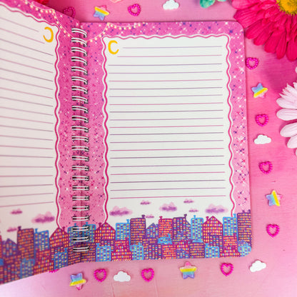 An open spiral-bound lined notebook with pink decorative borders and a colorful cityscape illustration along the bottom, styled on a pink background with flowers and small stickers.