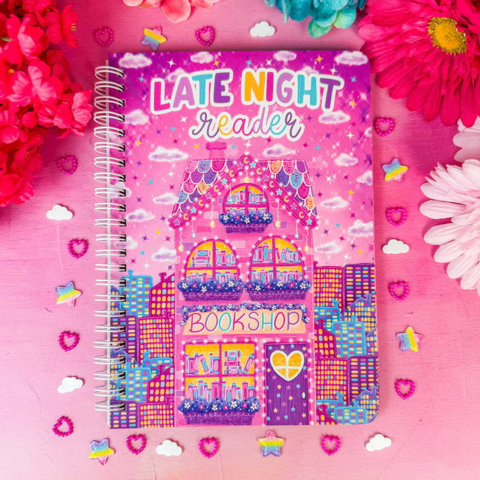 A spiral-bound notebook titled 'LATE NIGHT reader' with a colorful bookshop illustration on the cover, styled on a pink surface with flowers and small decorative stickers around it.