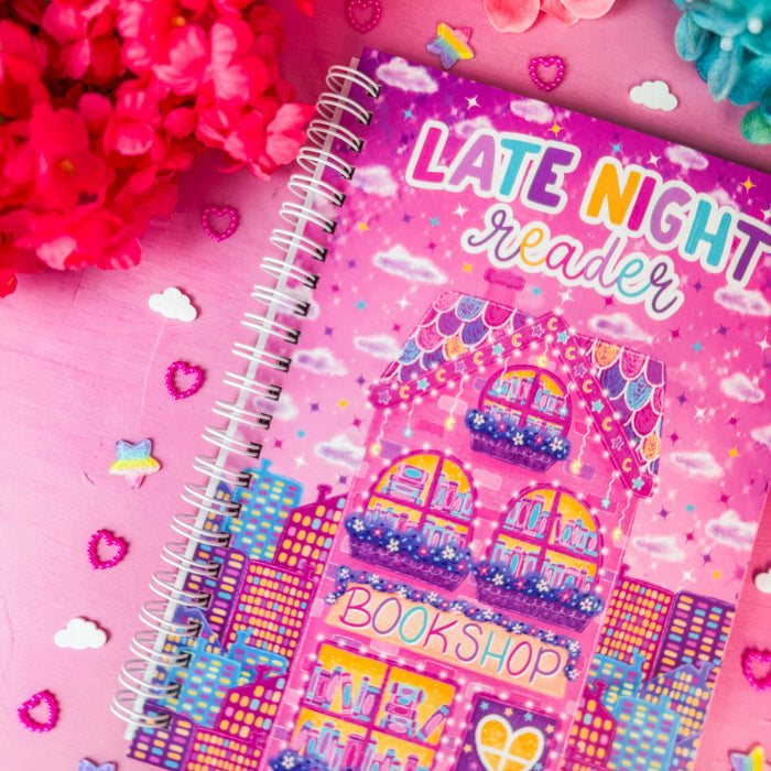 A pink spiral-bound notebook titled 'LATE NIGHT reader' with a colorful bookshop illustration on the cover, styled on a pink surface with bright flowers and small decorative hearts.