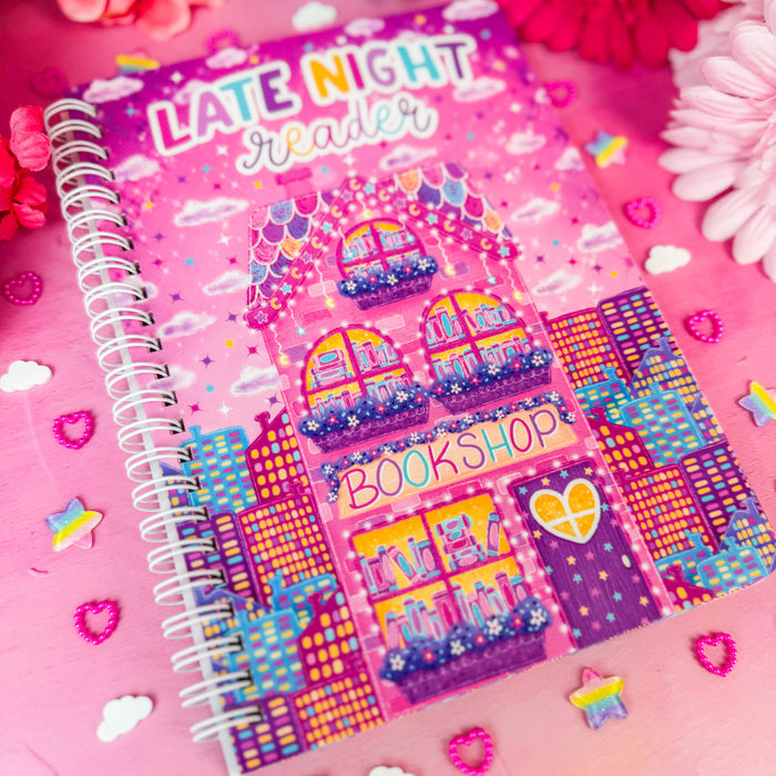 A pink spiral-bound notebook titled 'LATE NIGHT Reader' with a colorful illustration of a bookshop on the cover, styled on a pink surface with flowers and small decorative hearts.