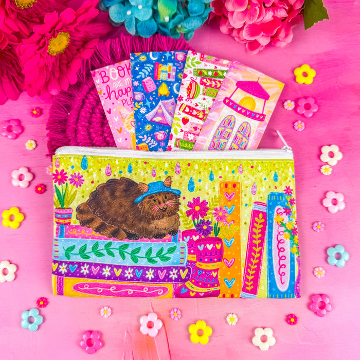 A bright, colorful zippered pouch illustrated with a fluffy brown cat, sitting on a stack of books, with vibrant flowers and books around it. Set against a pink background decorated with small flower shapes.