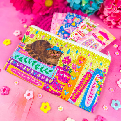 A colorful zippered pouch illustrated with a fluffy brown cat, sitting on a stack of books, with vibrant flowers and books around it. Set against a pink background decorated with small flower shapes.
