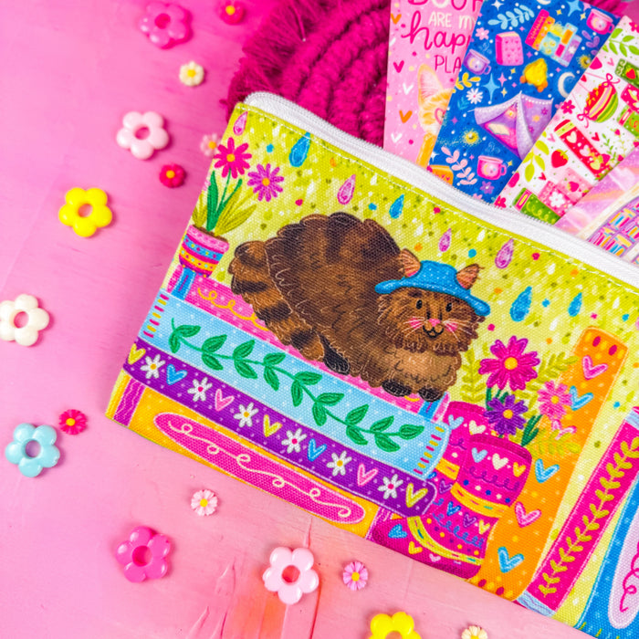 Close up of a colorful zippered pouch illustrated with a fluffy brown cat, sitting on a stack of books, with vibrant flowers and books around it. Set against a pink background decorated with small flower shapes.