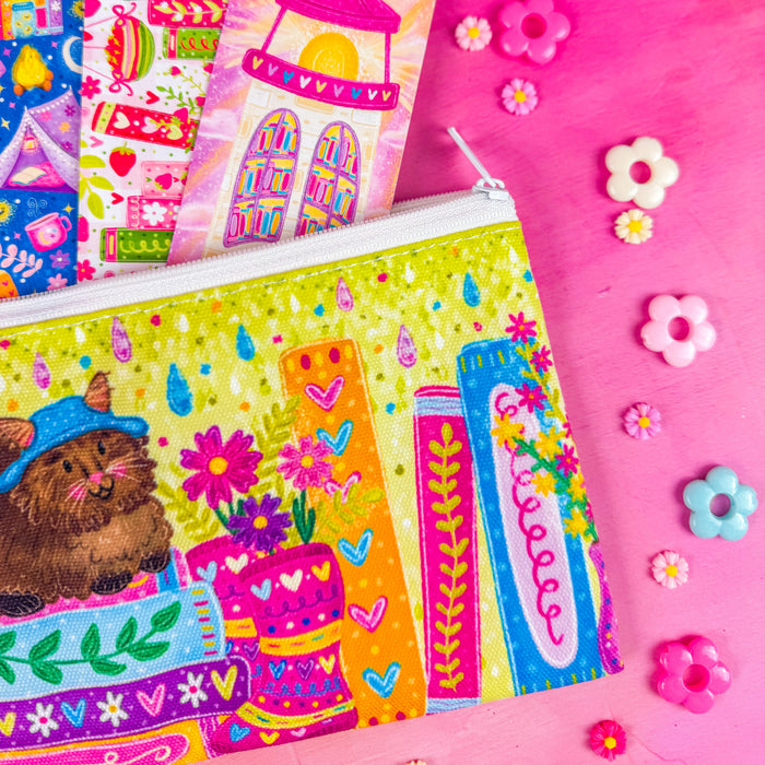 Close up of a colorful zippered pouch illustrated with a fluffy brown cat, sitting on a stack of books, with vibrant flowers and books around it. Set against a pink background decorated with small flower shapes.