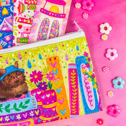 Close up of a colorful zippered pouch illustrated with a fluffy brown cat, sitting on a stack of books, with vibrant flowers and books around it. Set against a pink background decorated with small flower shapes.