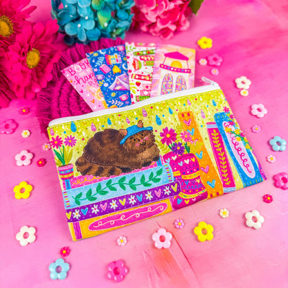 A colorful zippered pouch illustrated with a fluffy brown cat, sitting on a stack of books, with vibrant flowers and books around it. Set against a pink background decorated with small flower shapes.