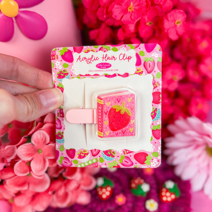 Acrylic hair clip with strawberry design held in front of pink flowers and a pink bag.