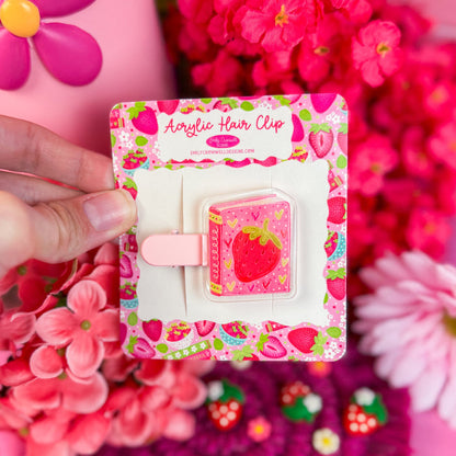 Acrylic hair clip with strawberry design held in front of pink flowers and a pink bag.