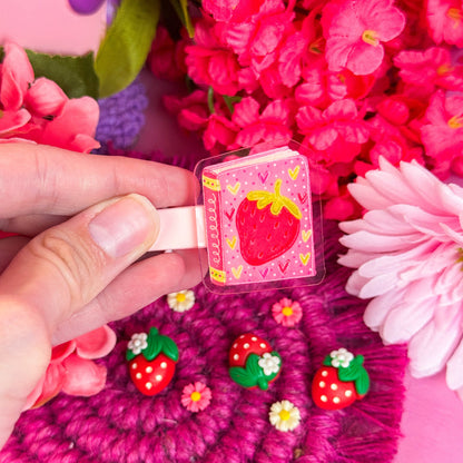 Hand holding a small hair clip with strawberry design on a pink textured surface with flowers.