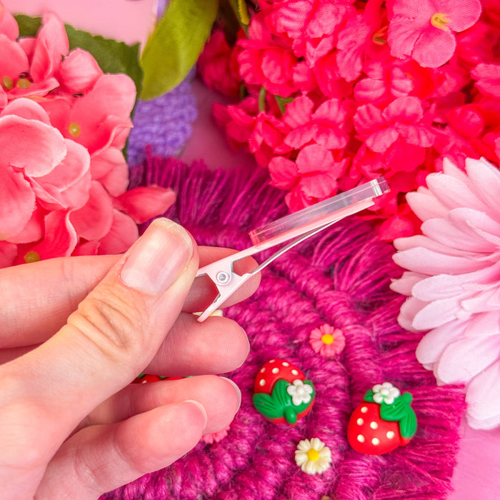 Hand holding a small hair clip with strawberry design against a colorful floral background