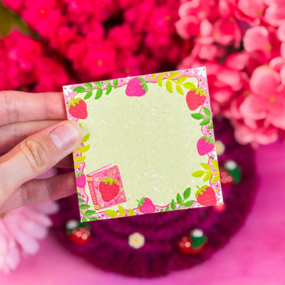 Hand holding a strawberry sticky note with pink flowers in the background