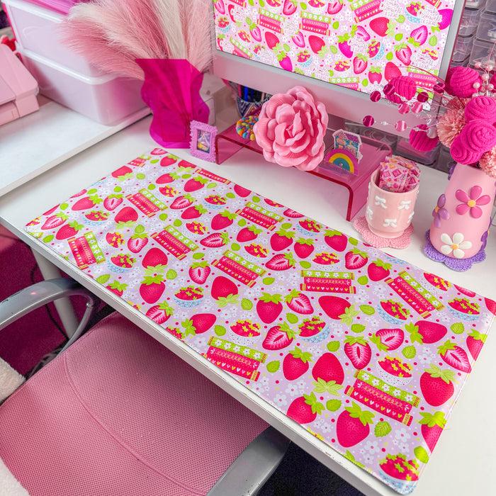 A pink and white strawberry-themed desk mat on a white desk, styled with a matching pink computer mousepad, a pink monitor, and various cute desk accessories.