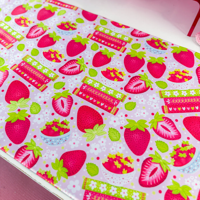 A pink and white strawberry-themed desk mat on a white desk, styled with a matching pink computer mousepad, a pink monitor, and various cute desk accessories.