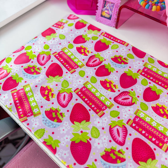 A pink and white strawberry-themed desk mat on a white desk, styled with a matching pink computer mousepad, a pink monitor, and various cute desk accessories.