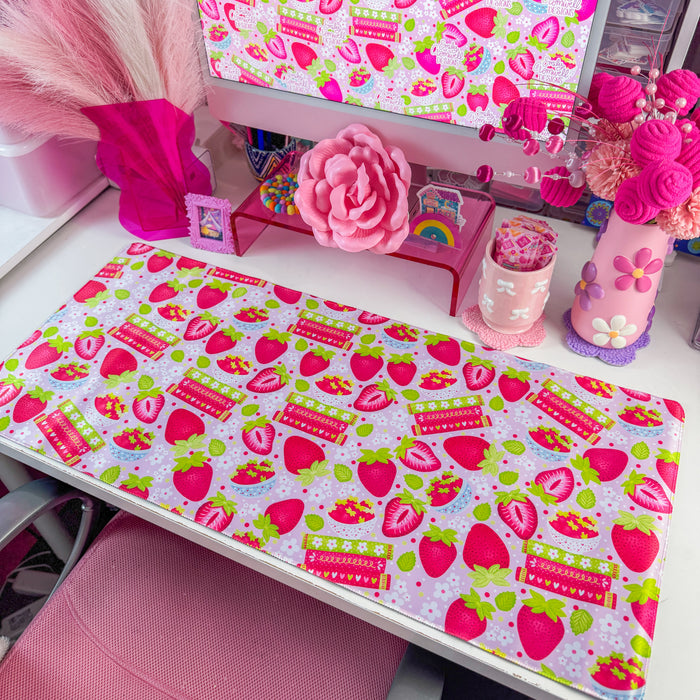 A pink and white strawberry-themed desk mat on a white desk, styled with a matching pink computer mousepad, a pink monitor, and various cute desk accessories.
